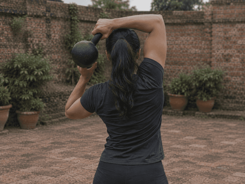 Seen from behind, a woman circles a kettlebell around her head, one hand on the handle and one under the bell