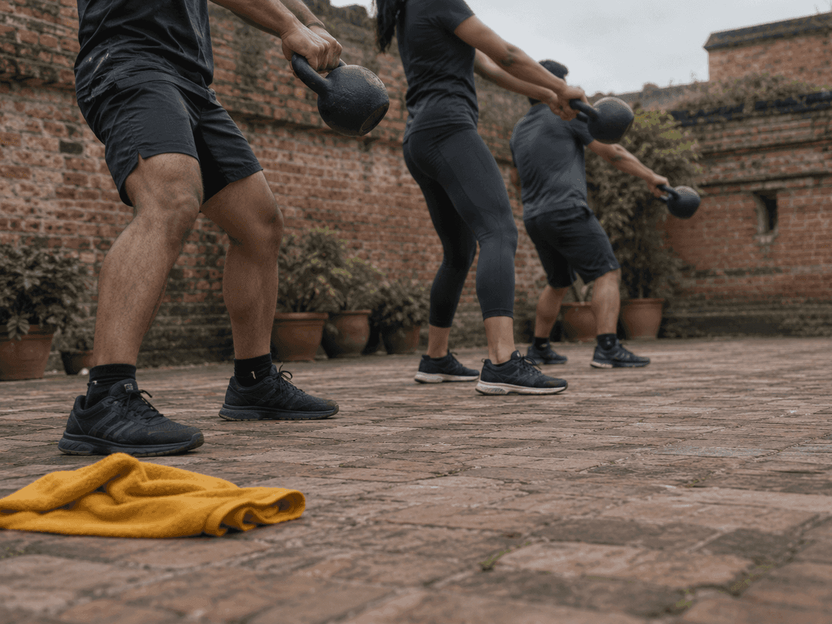 Three people swing kettlebells side by side in a brick-walled courtyard, a yellow towel on the paving in front